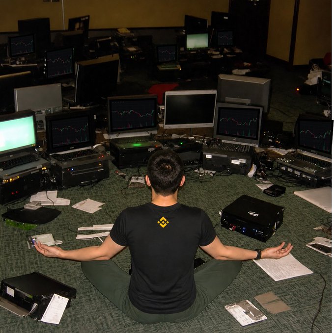 A man wearing a Binance-branded t-shirt sits cross-legged in a meditative pose surrounded by multiple computer monitors displaying trading charts and scattered papers.