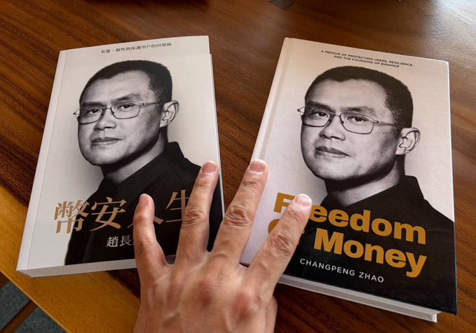 A hand gestures between two copies of Changpeng Zhao's book "Freedom Money"—the English edition on the right and its Chinese counterpart on the left—photographed on a wooden table, visually supporting the poster's appeal for a Vietnamese translation of CZ's memoir to be released and shared at a community meetup in Ho Chi Minh City.