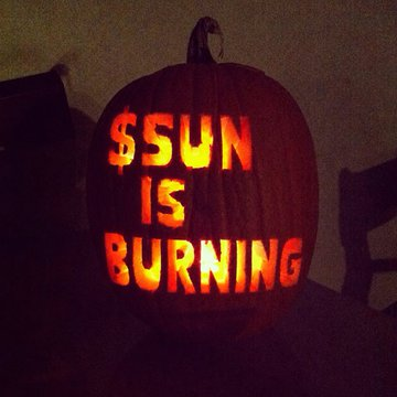 A glowing jack-o'-lantern carved with "$SUN IS BURNING" sits in the dark, cleverly referencing the Sun (SUN) token. The pumpkin's fiery glow pairs with the post's theme of powering borderless payments via Tron stablecoins, suggesting burning tokens as a deflationary mechanism to fuel the network's transaction ecosystem.