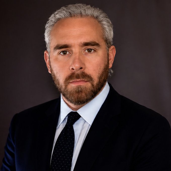 A professional portrait of a man with gray hair and a beard, wearing a dark suit, light blue shirt, and a dark polka-dot tie against a dark background.
