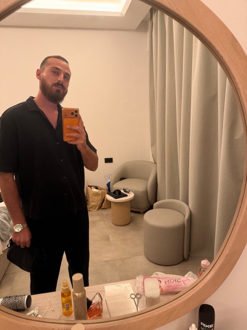 A bearded man in a black shirt takes a mirror selfie in a stylish dressing room with modern cream-colored seating, holding a smartphone with an orange case. Various toiletries, including Nuxe and AXE products, are scattered on the counter, suggesting a "Gm" (good morning) grooming routine as he prepares for the day.