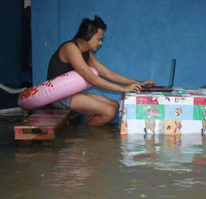 A young man wears headphones and works on a laptop while submerged in floodwater, using a pink swim ring for flotation and a table draped with cartoon-patterned cloth as his makeshift desk—embodying the meme's joke about staying "locked in" and focused on screens no matter how dire the circumstances.