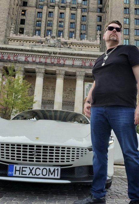 A man in a black t-shirt and sunglasses stands next to a white Ferrari with a 'HEX.COM1' license plate in front of a 'Palace of Culture' building.