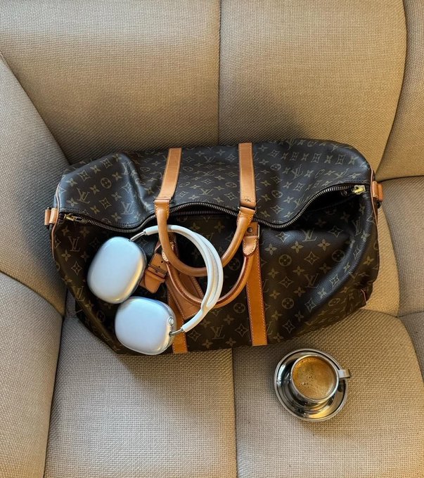 A Louis Vuitton monogram duffel bag sits on a beige couch with white over-ear headphones tucked inside it and a takeaway coffee cup resting on the seat beside it.