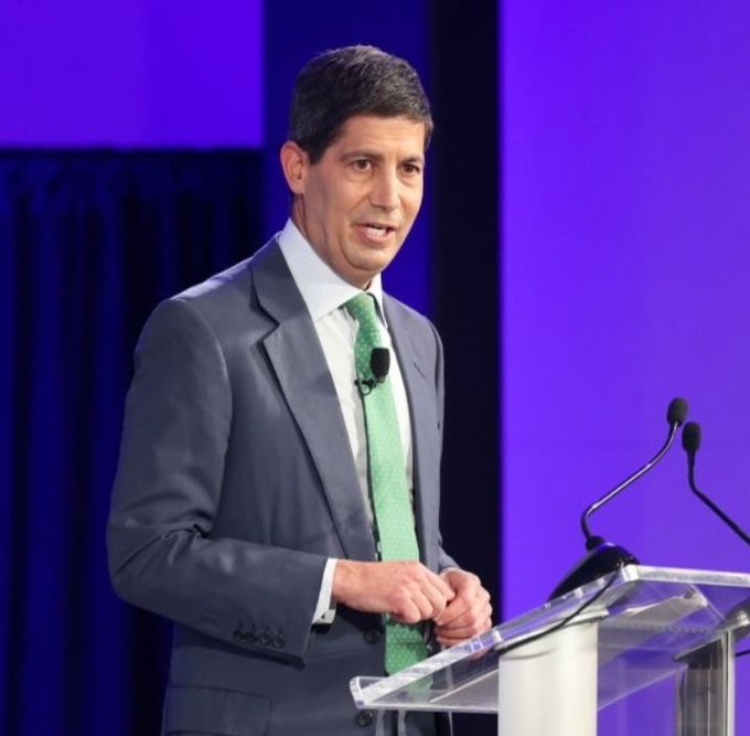 A man in a gray suit and green tie speaking at a clear podium with microphones against a blue curtain backdrop.