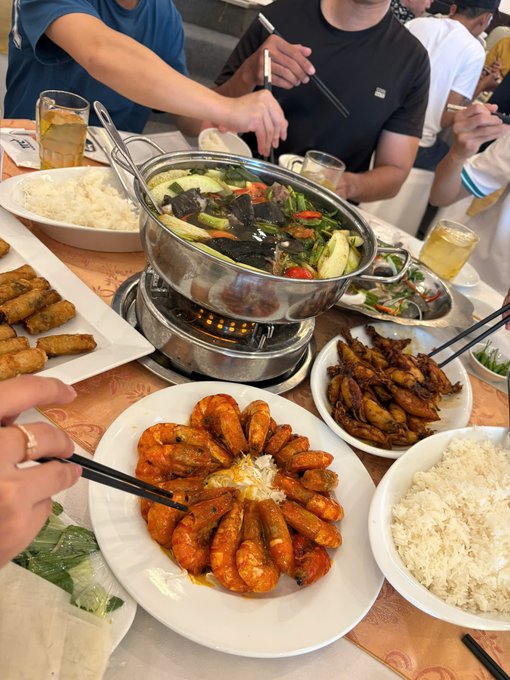 A table filled with a feast of Asian cuisine, featuring a central hot pot with vegetables, plates of white rice, fried spring rolls, glazed chicken wings, and a large platter of cooked shrimp arranged in a circle.