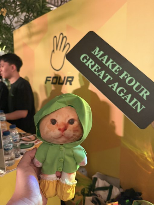 A small orange cat wearing a green hoodie and yellow pants being held up in front of a 'MAKE FOUR GREAT AGAIN' sign.