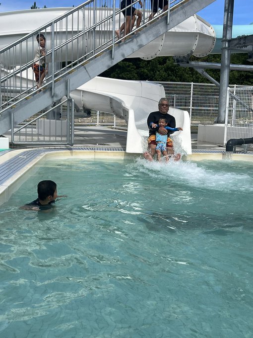 An adult and child slide down a white water slide into a pool, while another child swims nearby and two people stand at the top of the stairs.