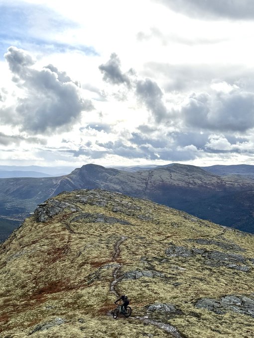 A scenic landscape photograph showing a mountain biker riding along a narrow rocky ridge under a partly cloudy sky.