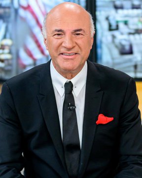 Kevin O'Leary, known as "Mr. Wonderful" from the TV show *Shark Tank*, is shown smiling in a professional portrait wearing a black suit with a black tie and a red pocket square, with a softly blurred studio background featuring an American flag. The image illustrates the accompanying post in which O'Leary, a prominent investor and crypto advocate, comments on the Clarity Act and his bold prediction that Bitcoin could reach $1 million if quantum computing challenges are resolved.
