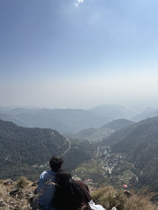 A scenic overlook where two people sit on a grassy hillside gazing down at a mountainous valley with winding roads and small villages below, under a hazy blue sky.