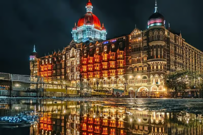 The Taj Mahal Palace Hotel in Mumbai illuminated at night, featuring a prominent red-lit dome and reflecting in water below.