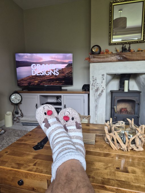 A first-person view of someone relaxing on a wooden coffee table wearing festive chicken-patterned socks, watching the TV show 'GRAND DESIGNS' with a fireplace visible in the background.