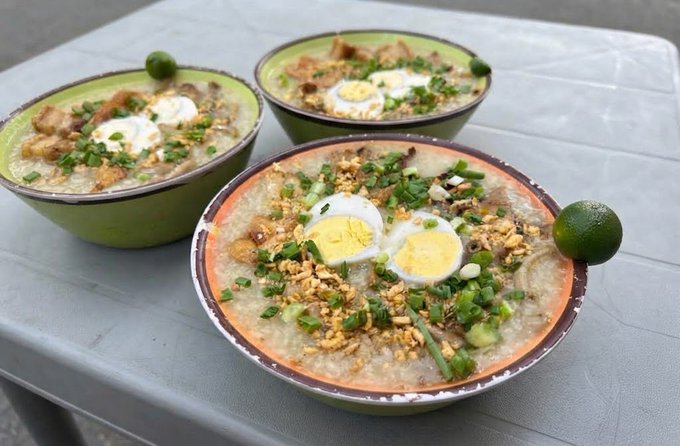 Three large bowls of Filipino-style lugaw or arroz caldo (rice porridge) topped with sliced boiled eggs, chives, crushed crackers, served on a gray table with calamansi limes.