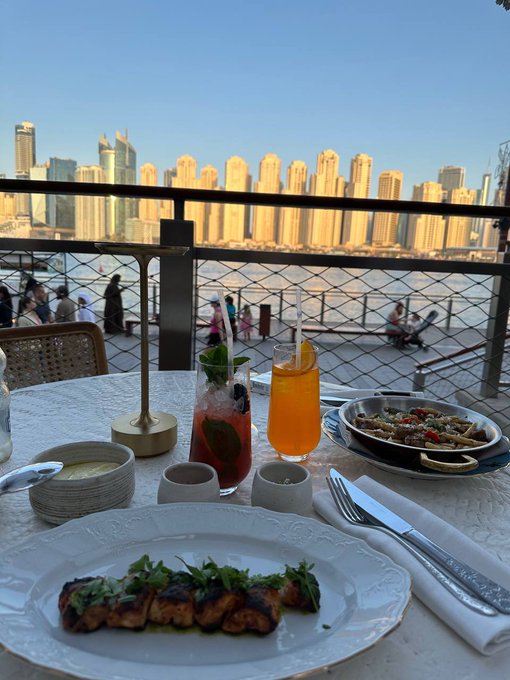 A dining table set with a plated meal, two drinks including what appears to be an Aperol spritz, and a view of a city skyline across water in the background.
