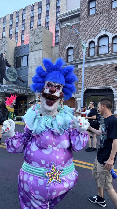 A creepy clown with bright blue hair, white face paint, and a purple polka-dot costume stands on a city street in front of brick buildings.