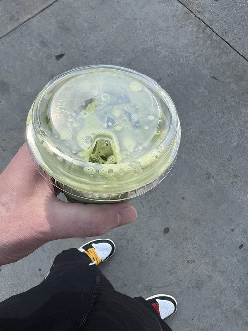 A first-person view of a hand holding a clear plastic cup of iced green-colored beverage with a lid and straw, standing on a concrete sidewalk.