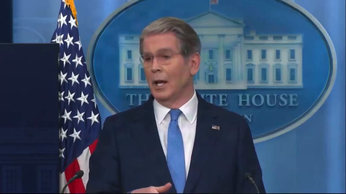 The image shows a man wearing glasses, standing at a podium with his hands on his hips. He appears to be giving a speech or addressing an audience. The setting seems to be indoors, possibly within a room that has a large American flag displayed prominently in front of him. The man is dressed formally, suggesting the event might be significant or formal in nature.