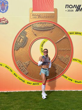 A woman in a sporty outfit posing in front of a large circular medal prop with Russian text including 'ПОЛУМАРАФОН' and '2026' visible on the backdrop.