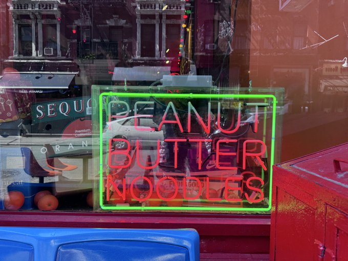 A storefront window displays a neon sign reading 'PEANUT BUTTER NOODLES' in red lettering outlined in green, with reflections of city buildings and street activity visible in the glass.