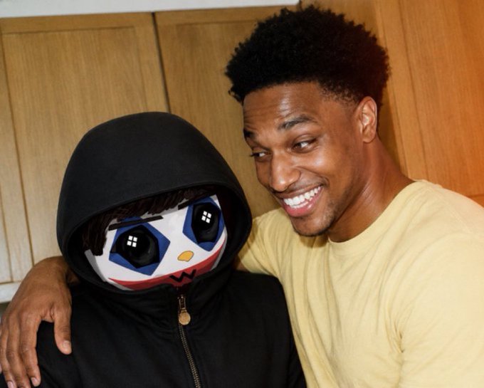 A man in a cream-colored t-shirt smiles broadly while posing with an arm around a person in a black hoodie wearing an oversized cartoonish mask with large expressive eyes. The image relates to the post by visually representing the "$ANSEM" collaboration, as the masked figure appears to be the memecoin's mascot.