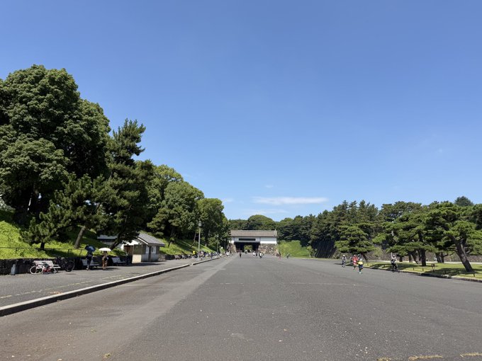 A wide paved path lined with green trees leads toward a traditional Japanese gate structure under a clear blue sky, with a few people walking in the distance.