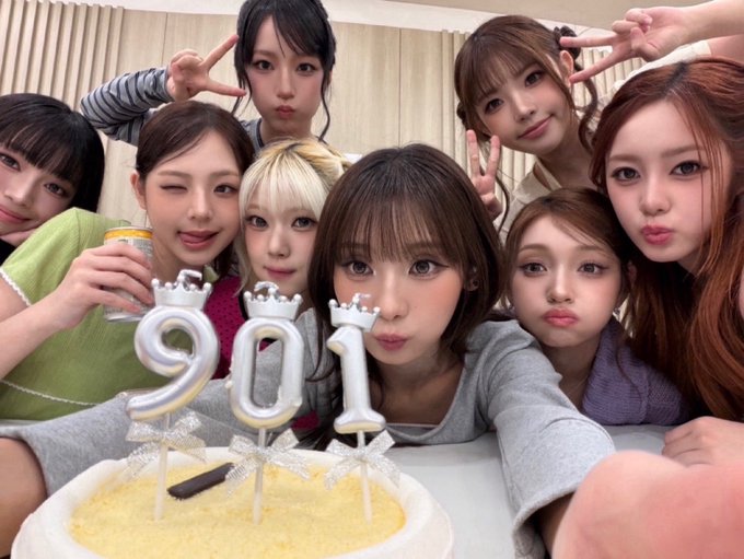 A group of nine young women posing together behind a birthday cake decorated with '201' shaped candles and small crown toppers.