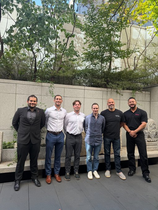 A group photo of seven people standing together outdoors against a stone wall with greenery behind them, dressed in business casual and smart attire.