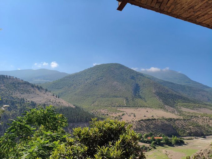 A scenic landscape photograph showing green forested mountains under a clear blue sky, with a portion of a thatched roof visible in the upper right corner and green foliage in the foreground.