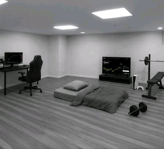 A minimalist bedroom setup features a bed, desk with a computer monitor, TV displaying a stock chart, weight bench with a dumbbell, and a PlayStation 5 console, all in a gray-toned room.