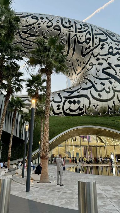 The Museum of the Future in Dubai, featuring its distinctive torus-shaped structure covered in Arabic calligraphy, with palm trees and visitors in the foreground.