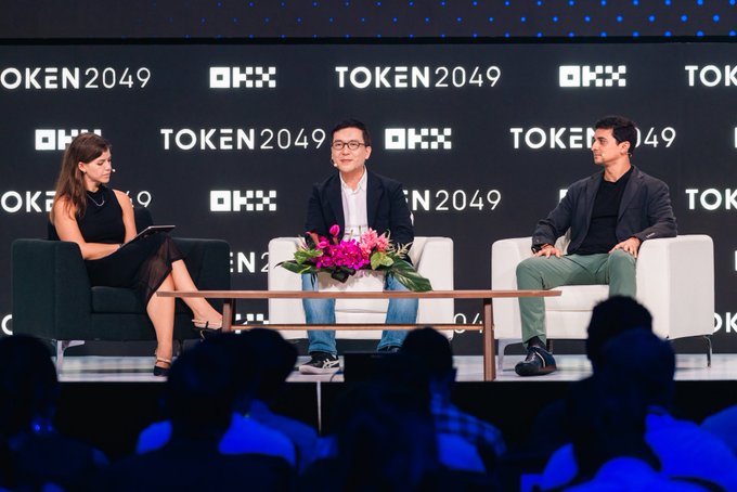 A moderator interviews two panelists at the OKX-hosted TOKEN2049 conference, with the event's branding displayed prominently across the backdrop, illustrating the crypto industry's push to bring blockchain technology—like Solana—to mainstream adoption and "a billion pockets."