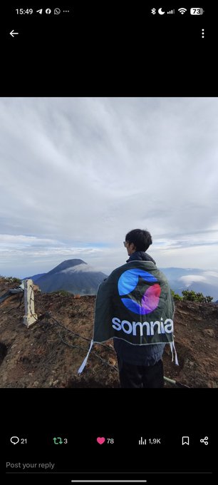 A person stands on a mountain summit wearing a 'somnia' branded jacket, with a phone screenshot showing 21 comments, 3 retweets, 78 likes, and 1.9K views.