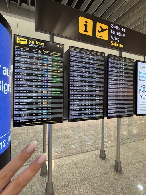 The image shows a person with their hand outstretched, likely posing for a photo at an airport terminal. In front of them, there are three large signs displaying flight information such as departure times, arrival times, and destinations. The signs are positioned on poles, providing easy visibility from various angles. The background features the interior of an airport terminal, complete with seating areas and other amenities typically found in such spaces.