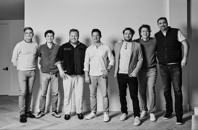 The image features a group of six men standing next to each other, posing for a photo. They are all dressed in casual attire with some wearing ties as well. The background appears to be a wall or a large piece of furniture that serves as their backdrop. The photograph is black and white, giving it an artistic touch. The image also contains text that reads "Building something great". This suggests that the group might be involved in a project or endeavor related to building or creating something significant.