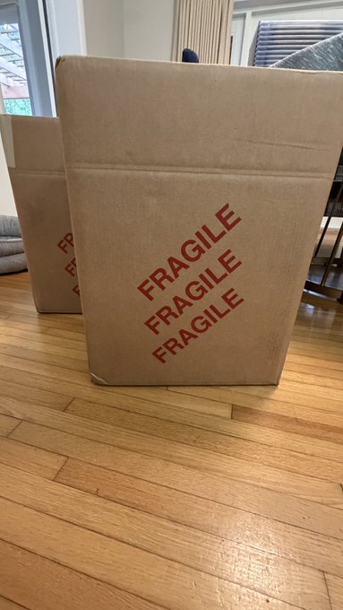 A large cardboard box sits on a hardwood floor, prominently displaying "FRAGILE FRAGILE FRAGILE" in bold red lettering, with another similar box partially visible behind it—visually representing one of the three big boxes of graded VeeFriends comics just delivered from CGC.