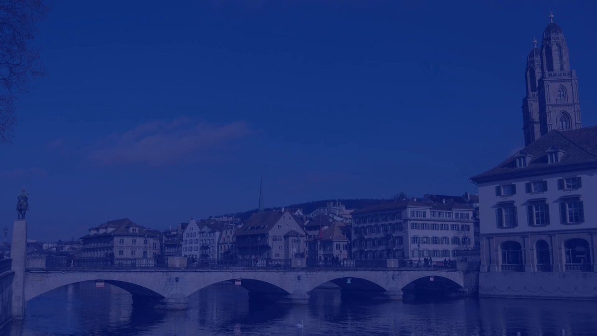The image shows a view of a cityscape with a river running through it, featuring a bridge over the water. The sky above appears blue, suggesting that it might be early morning or late afternoon when the sun's position is lower on the horizon. In the background, there are buildings and a church, which adds to the overall atmosphere of the scene. The image does not contain any text, so we cannot determine if this is an advertisement for a product or event related to finance.