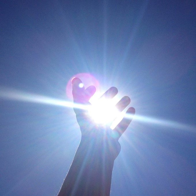 A silhouetted hand reaching up against a bright blue sky, with the sun shining through the fingers creating a lens flare effect.
