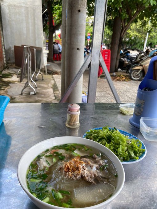 A bowl of Vietnamese noodle soup (pho) topped with fried shallots and greens, served on a metal table alongside a plate of fresh herbs. The view looks out onto a street where parked scooters and people sitting at other food stalls are visible.