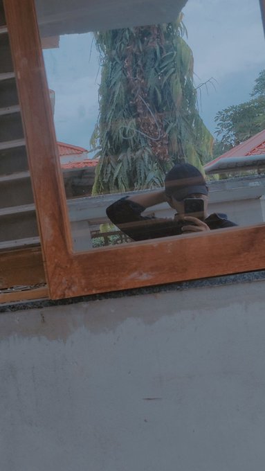 A mirror reflection showing a person wearing a black cap taking a selfie with a smartphone near an open window with plants hanging outside.