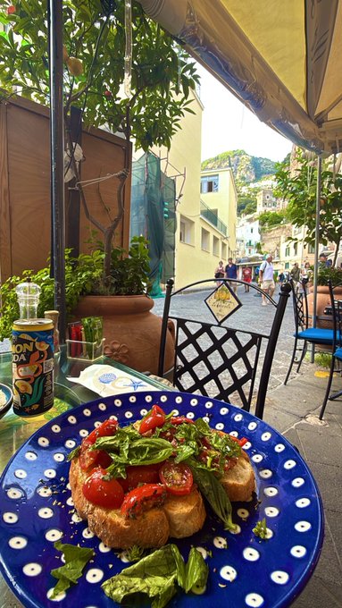 A vibrant Mediterranean-style bruschetta topped with tomatoes and fresh basil served on a blue and white polka-dot plate at an outdoor cafe, with a can of Longrider beer and a charming street view in the background.