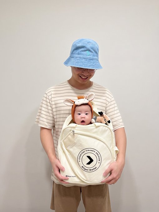 A person wearing a striped shirt and blue bucket hat holds a baby in a deer-themed carrier against their chest; the carrier features a logo reading 'A BETTER FINANCE WORLD' and 'Bright Wallet'.