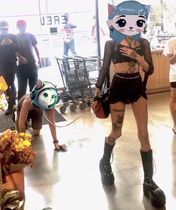 Two people in a shopping area wear cartoon cat filters over their faces, with one crawling on the floor on a leash and the other standing in a black mesh outfit with platform boots and visible tattoos.