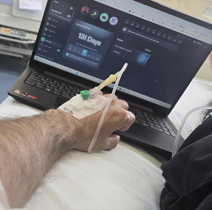 A patient's hand with an IV line rests on a hospital bed while using a laptop displaying a streak counter showing '131 Days' and 'Upvote Streak'.