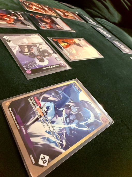 A close-up view of a tabletop card game in progress on a green cloth surface, featuring Azuki TCG cards including a "Robin" character card with "20" damage and a "Home Predator" card, with several other cards laid out in play. The image captures the user's first hands-on experience with the newly launched Azuki TCG, played casually at home since no official European stores carry the game yet.