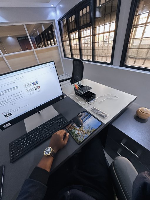 A first-person view of someone working at a desk setup, with one hand on a mouse displaying a nature scene, facing a monitor showing a webpage.