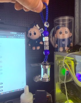 A hand holds a blue beaded lanyard with a small glass vial charm in front of a computer monitor, with plush figures and a PC case visible in the background.