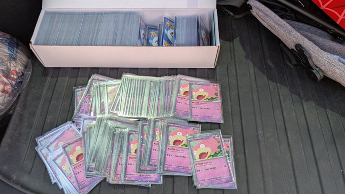 A collection of Pokémon trading cards spread out on a dark surface, with a white box containing more cards in the background; the cards appear to feature Slowbro illustrations.