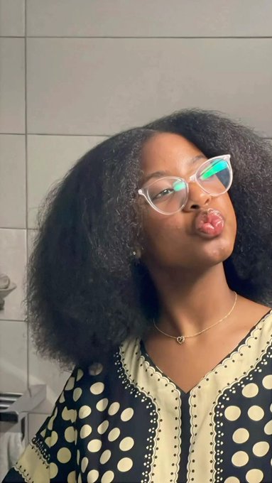 A selfie of a woman with glasses making a playful pouting expression, wearing a polka dot top and a heart necklace.