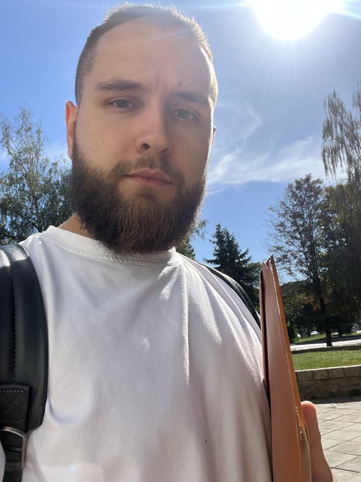 An outdoor selfie of a bearded man wearing a white t-shirt and carrying a backpack, with a brown folder under his arm on a sunny day.